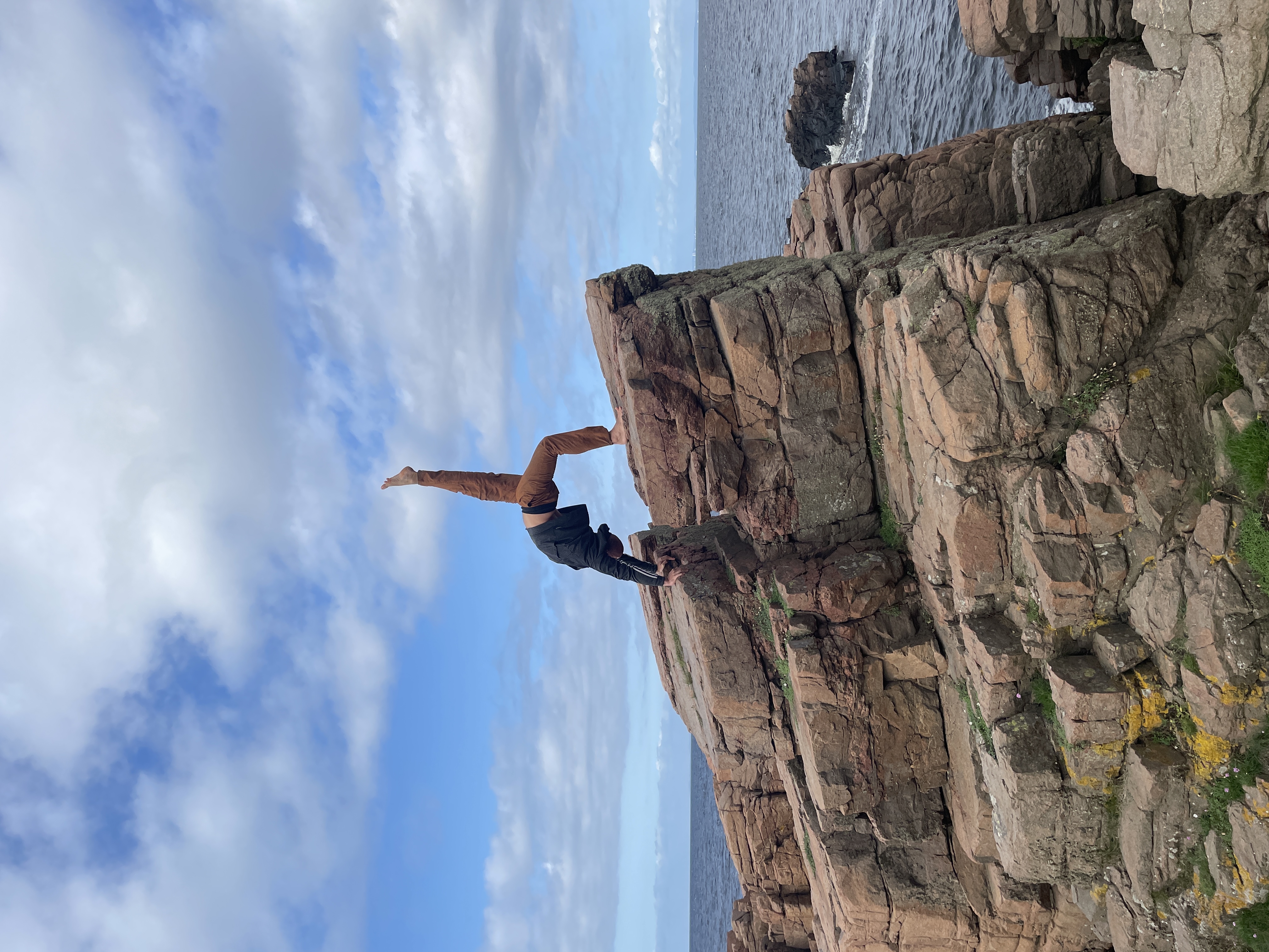 The yoga teacher does a backbend pose with one leg pointed to the sky, atop a high rocky outcropping by the ocean.